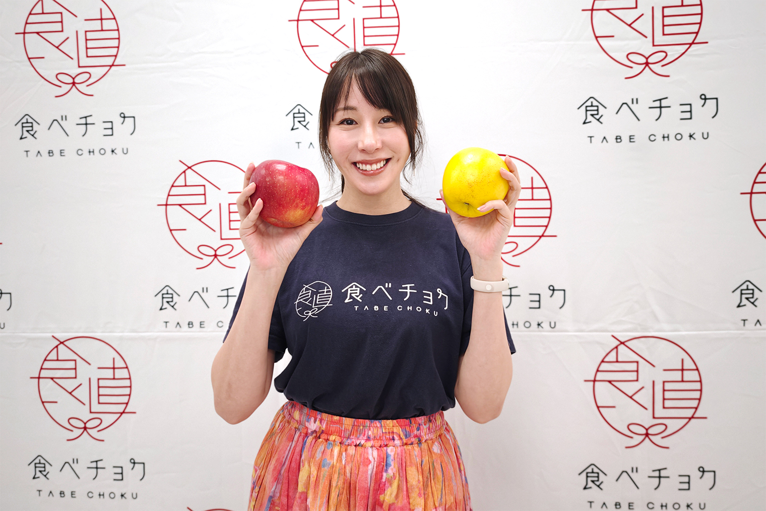 食べチョクのタペストリーを背景に、2色のりんごを手にして笑顔の秋元さん